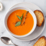 Bowl of Instant Pot creamy tomato soup next to a spoon and slice of baguette.