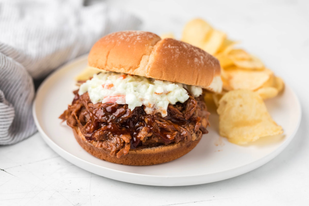 Barbecue beef sandwich topped with coleslaw next to potato chips. 